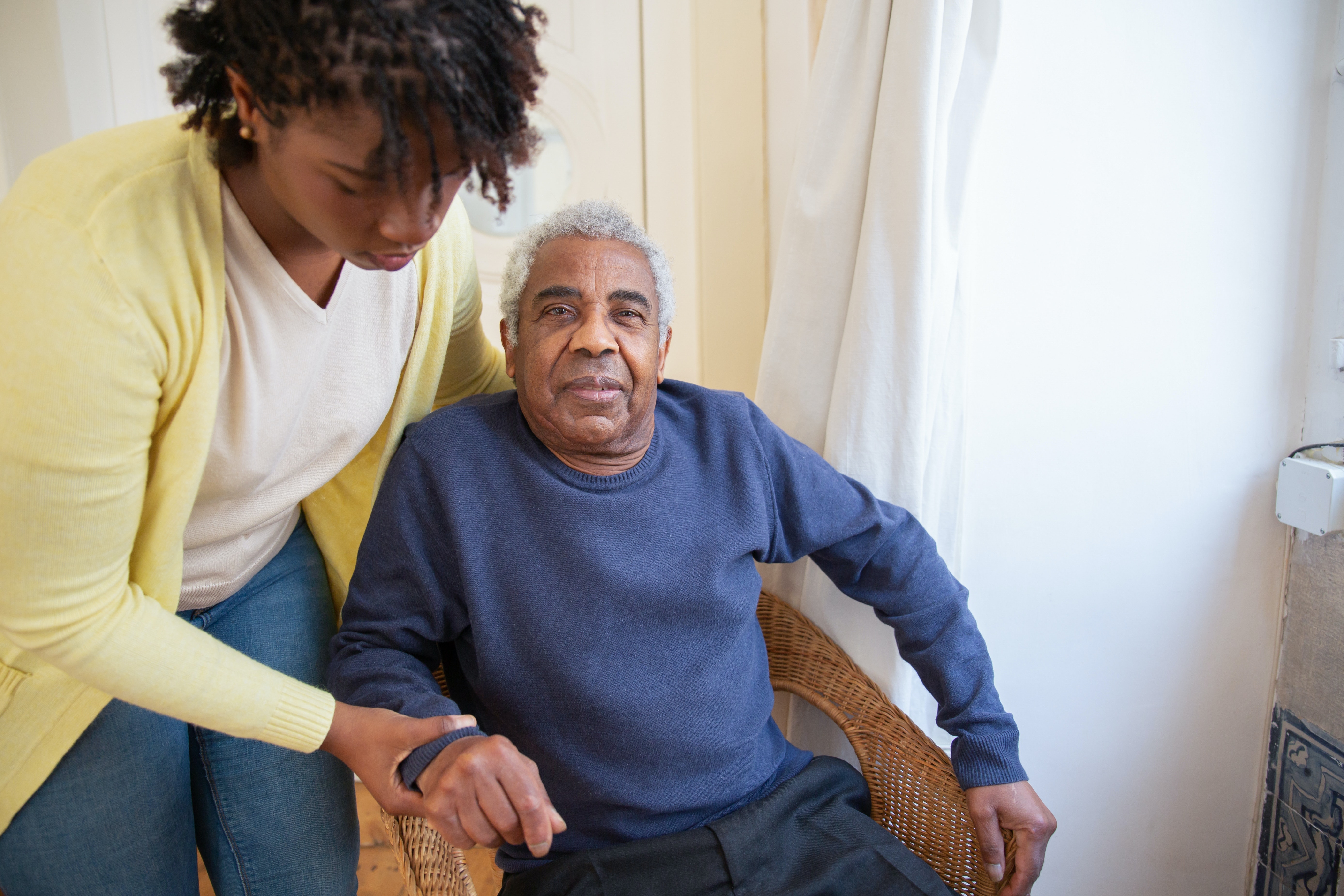 A female caregiver wearing a yellow sweater, helping a senior client to sit properly in his chair. He’s wearing a blue sweater and smiling.